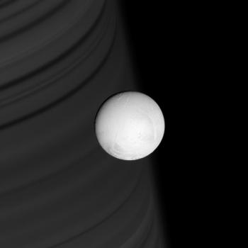 Ring shadows line the face of distant Saturn, providing a backdrop for the brilliant, white sphere of Enceladus