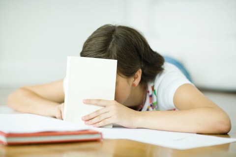 Teen girl lying on floor, doing homework, holding book in front of face